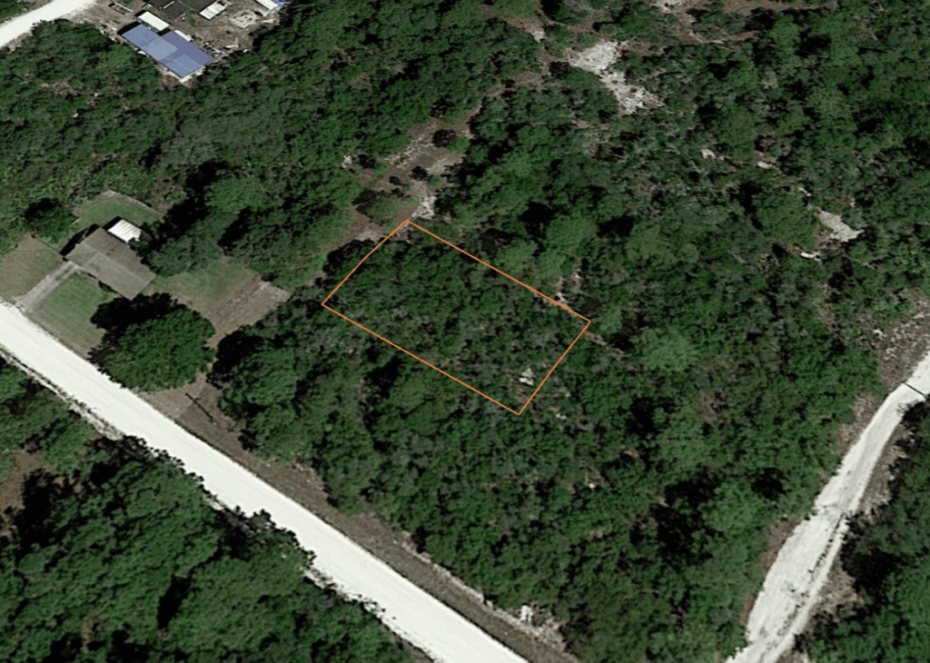 Aerial view of a rectangular plot outlined in orange within a forested residential area, with a road at the bottom and a house to the left.