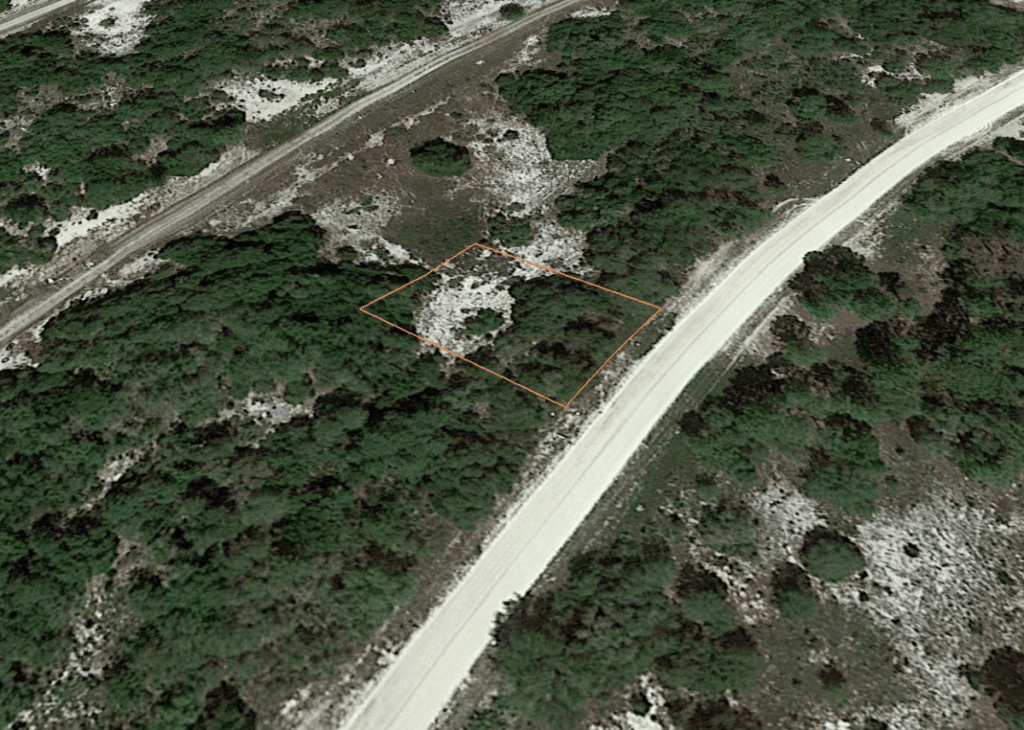 Aerial view of a rectangular plot outlined in orange beside a dirt road, surrounded by sparse trees and rocky ground.