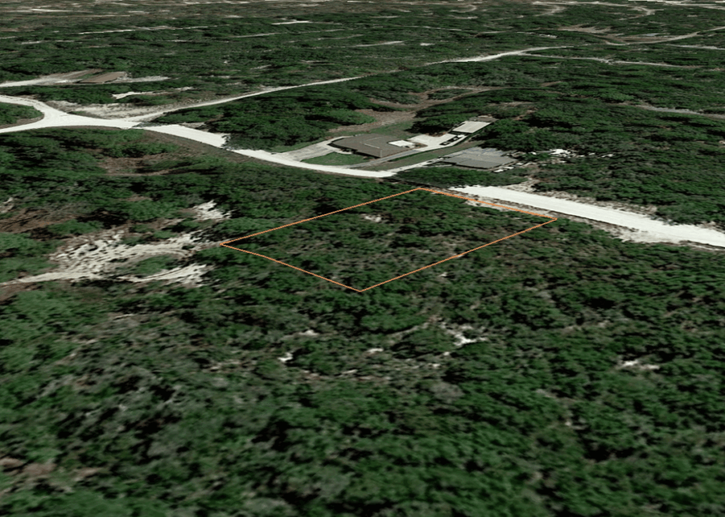 Aerial view of a rectangular wooded plot outlined in orange beside a dirt road; nearby buildings hint at development.