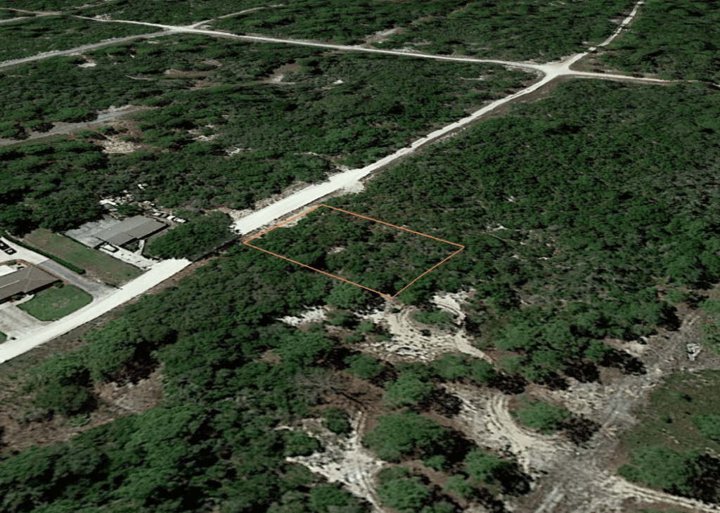 Aerial view of a rectangular lot outlined in orange among trees, adjacent to a dirt road near a residential area.