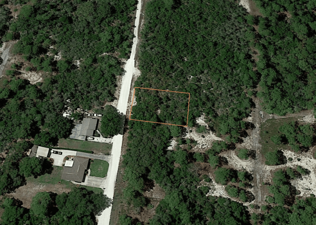 Aerial view of a forested residential area with an orange-outlined rectangular lot beside a light road.