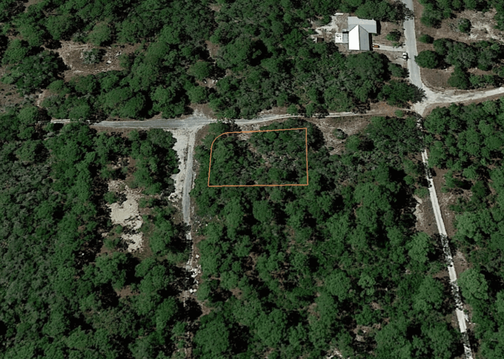 Aerial view of a forested area with an orange rectangle marking a vacant lot between dirt roads near a small building at the top edge of the image.