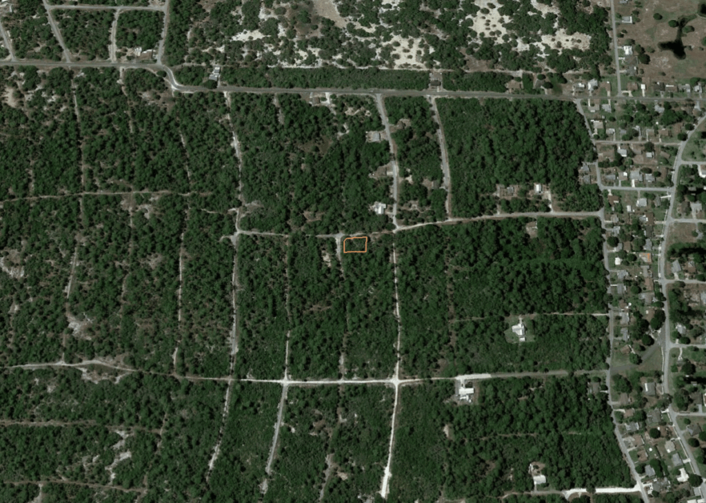 Aerial view of a forested area with a small rectangular plot outlined in orange at center-left, surrounded by trees and roads, near a residential area to the right.