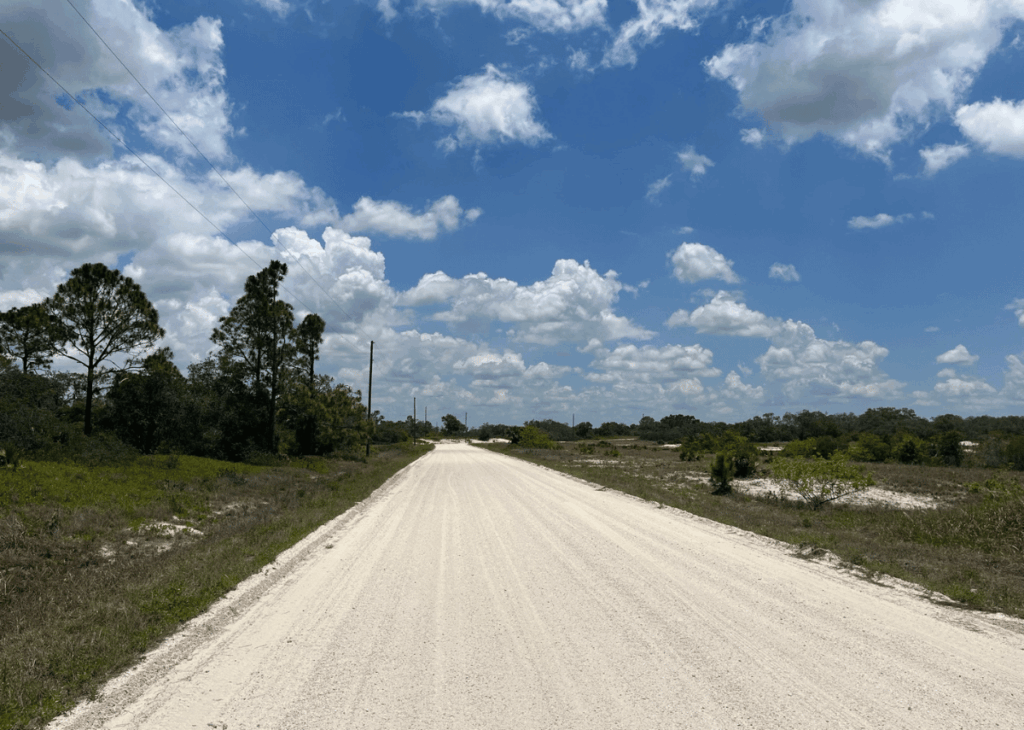 Dusty white dirt road stretches into the distance under a bright blue sky with scattered clouds, flanked by sparse trees and brush on both sides with power lines on the left.
