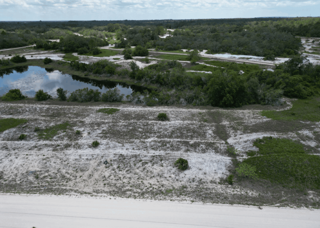 Aerial view of a sandy, scrubby landscape with a small winding pond and scattered bushes, under a partly cloudy sky.