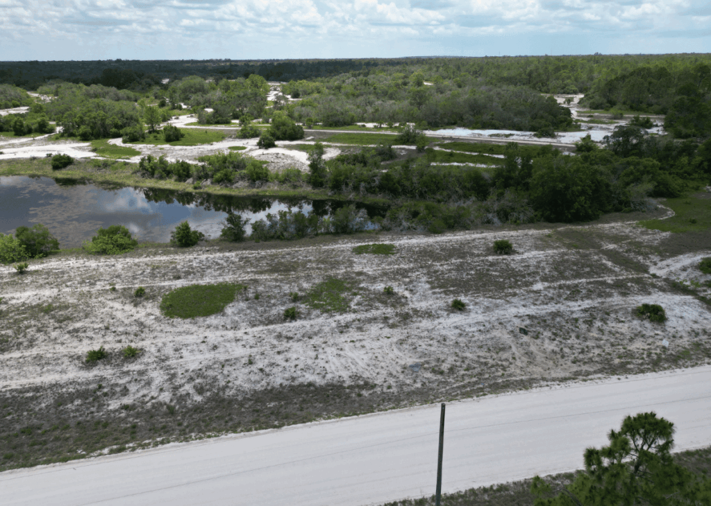 Open, undeveloped lot with sandy soil and sparse shrubs beside a paved road; a waterway and tree line appear in the distance under a cloudy sky.