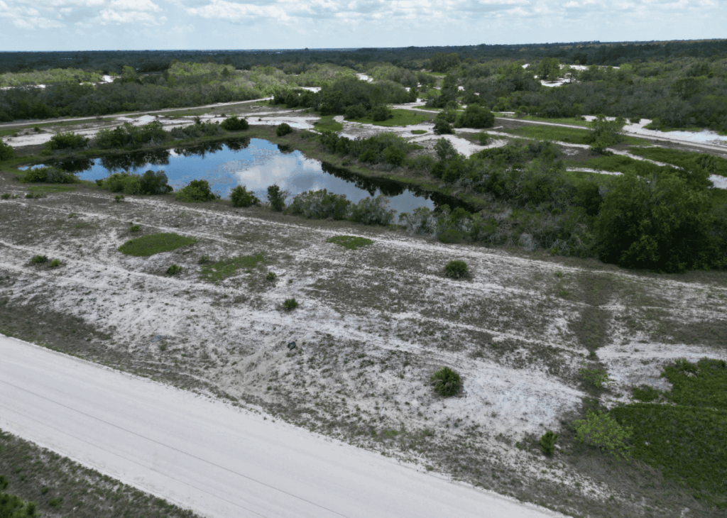 Aerial view of a sandy landscape with a small irregular pond surrounded by shrubs and trees; a dirt road runs along the bottom edge.